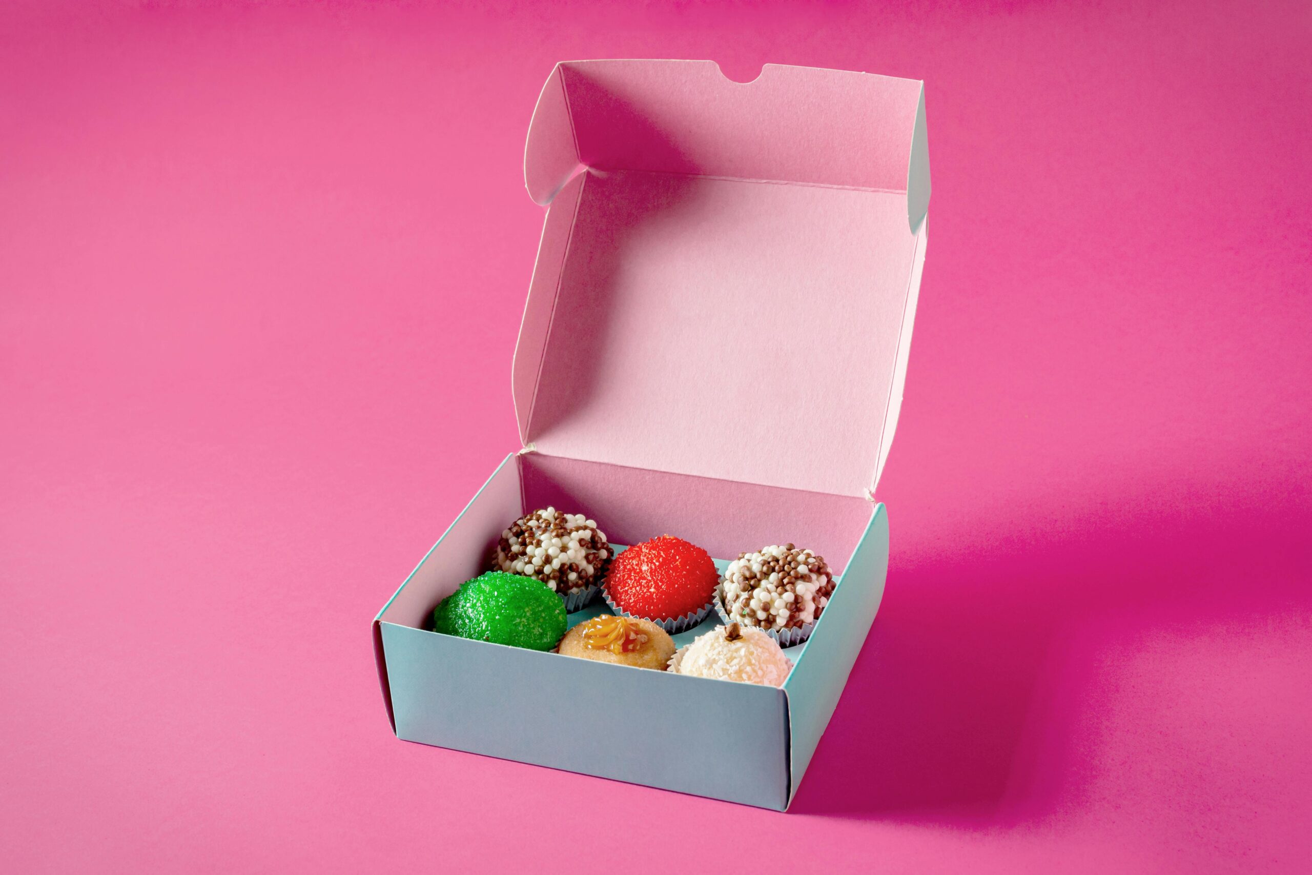 An open box of assorted cupcakes on a vibrant pink background, highlighting the colorful toppings.
